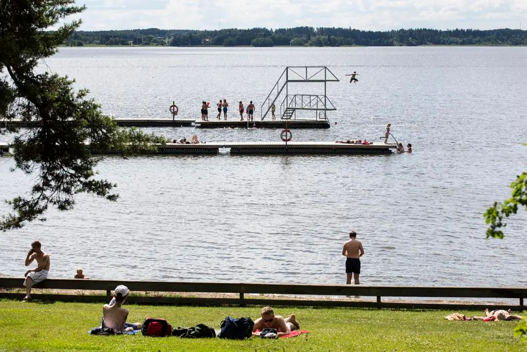 Strand med gräs och sand. I förgrunden syns badande barn och vuxna i vattnet och på bryggan.