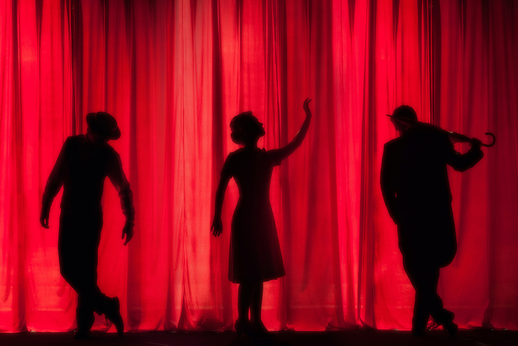 Bild på silhuetten av tre personer som står framför ett rött draperi på en scen.