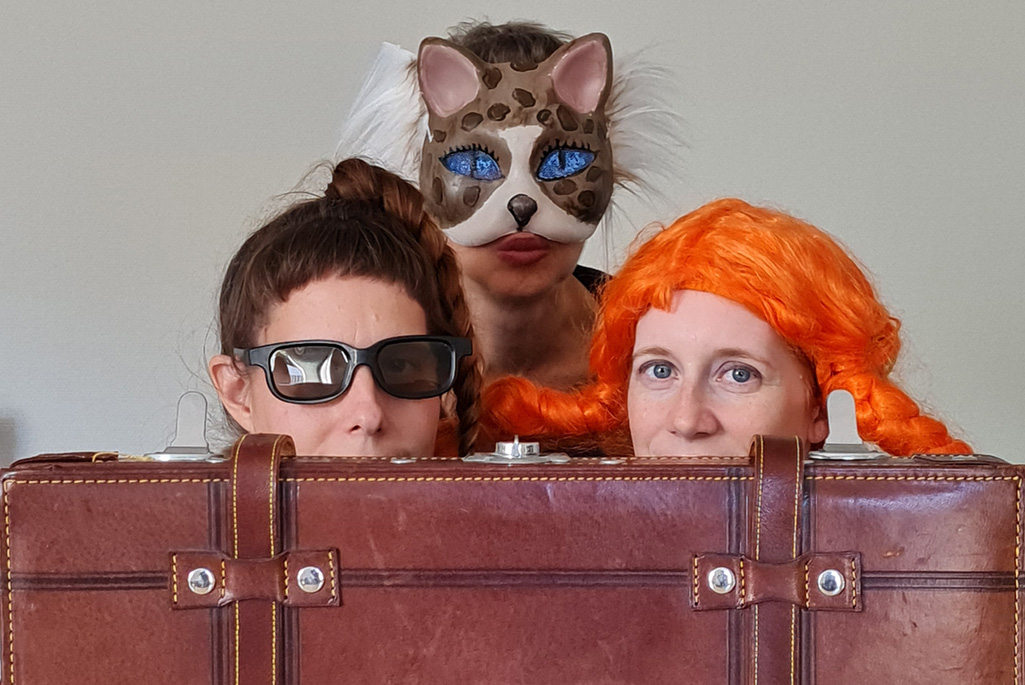 Bild på tre personer i maskeradkläder bakom en koffert som tittar fram.
