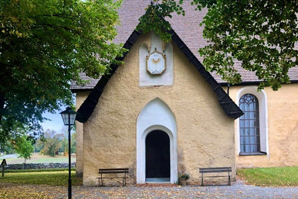 Bild på Veckholms kyrka. En gulaktig stenbyggnad med vita detaljer runt dörr och fönster.